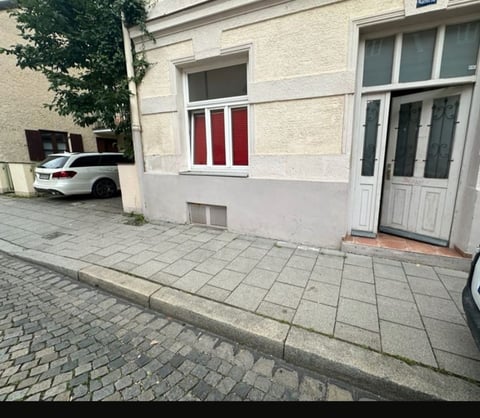 Residential building exterior with white facade, red-striped window, wooden door, paved courtyard, and parked white car