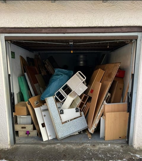 Crowded garage filled with stacked wooden boards, cardboard boxes, a step ladder, and miscellaneous stored items