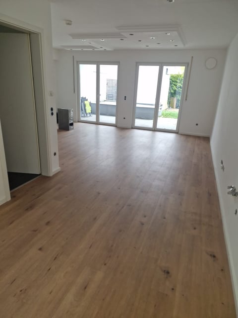 Empty modern living room with wood flooring, white walls, recessed lighting, and glass doors leading to an outdoor patio area
