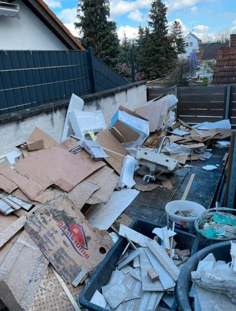 Cluttered construction waste and debris piled in an outdoor dumpster area with blue metal fencing and evergreen trees visible in background