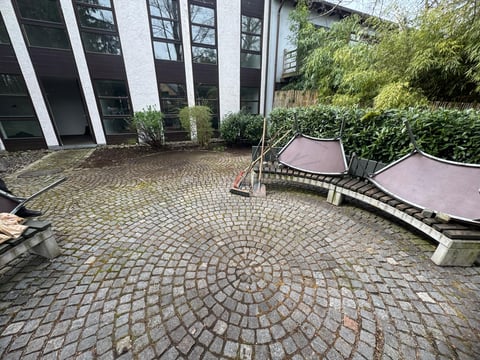 Outdoor courtyard with circular brick paving, modern building with white columns, green hedges, and purple bench seating
