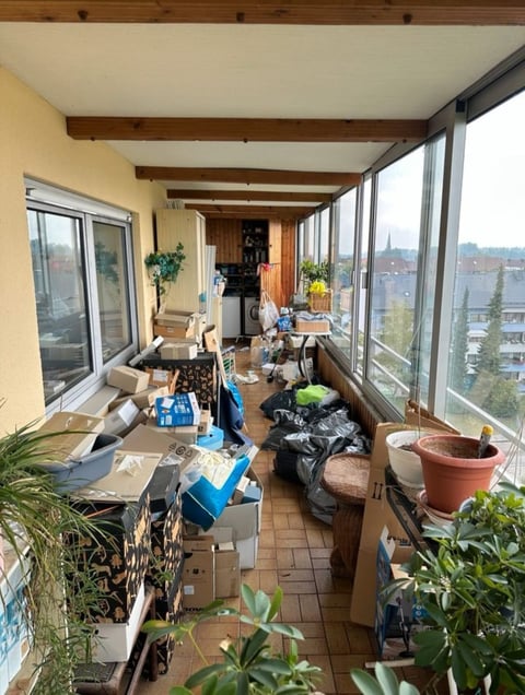 Long glass-enclosed balcony filled with potted plants, storage boxes, and household items, overlooking a cityscape with water in the distance