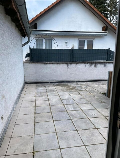 Residential courtyard with gray tile patio, dark metal balcony railing, and white-walled house with windows