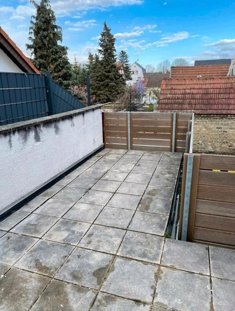 A residential rooftop terrace with stone paving tiles, wooden railings, and overlooking a neighborhood with brick-roofed houses and evergreen trees on a clear day