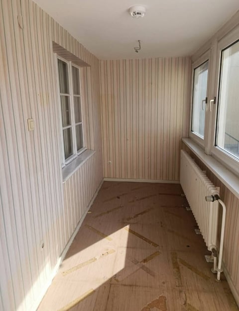 Empty room with beige vertical striped wallpaper, wooden floor, two windows, radiator, and a door along the walls